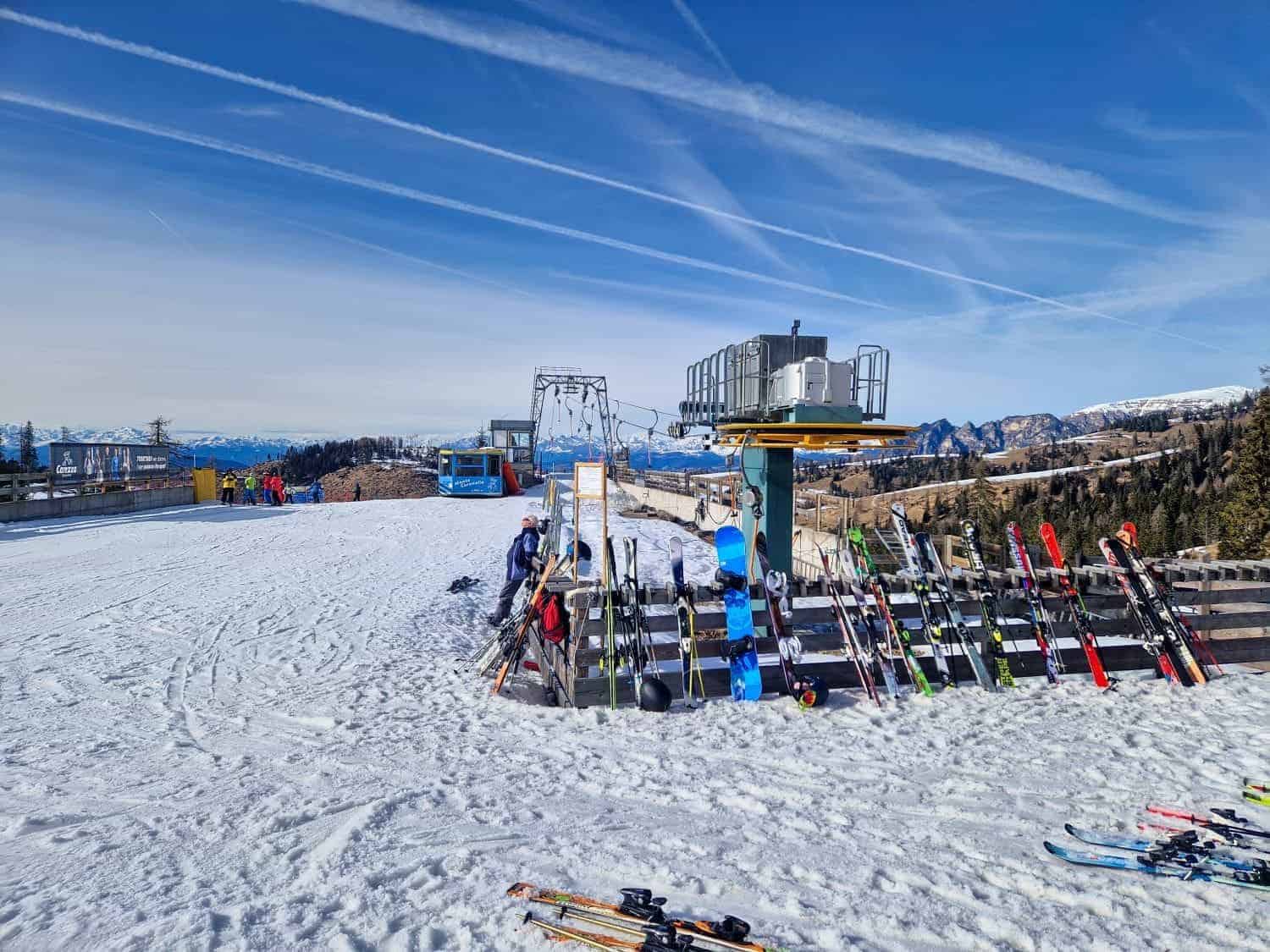 A snowy ski slope with several pairs of skis and poles lined up near a ski lift. Several people are preparing to ski, and a cable car is visible in the background under a clear blue sky with streaks of clouds.
