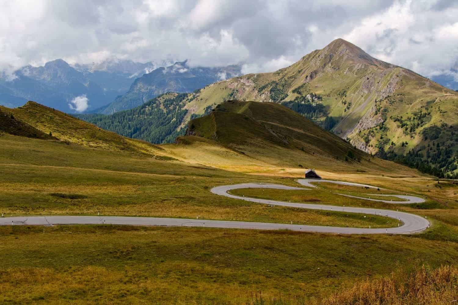 A winding road curves through a lush green valley with mountains in the background under a cloudy sky. A small building stands near the road surrounded by grassy hills.