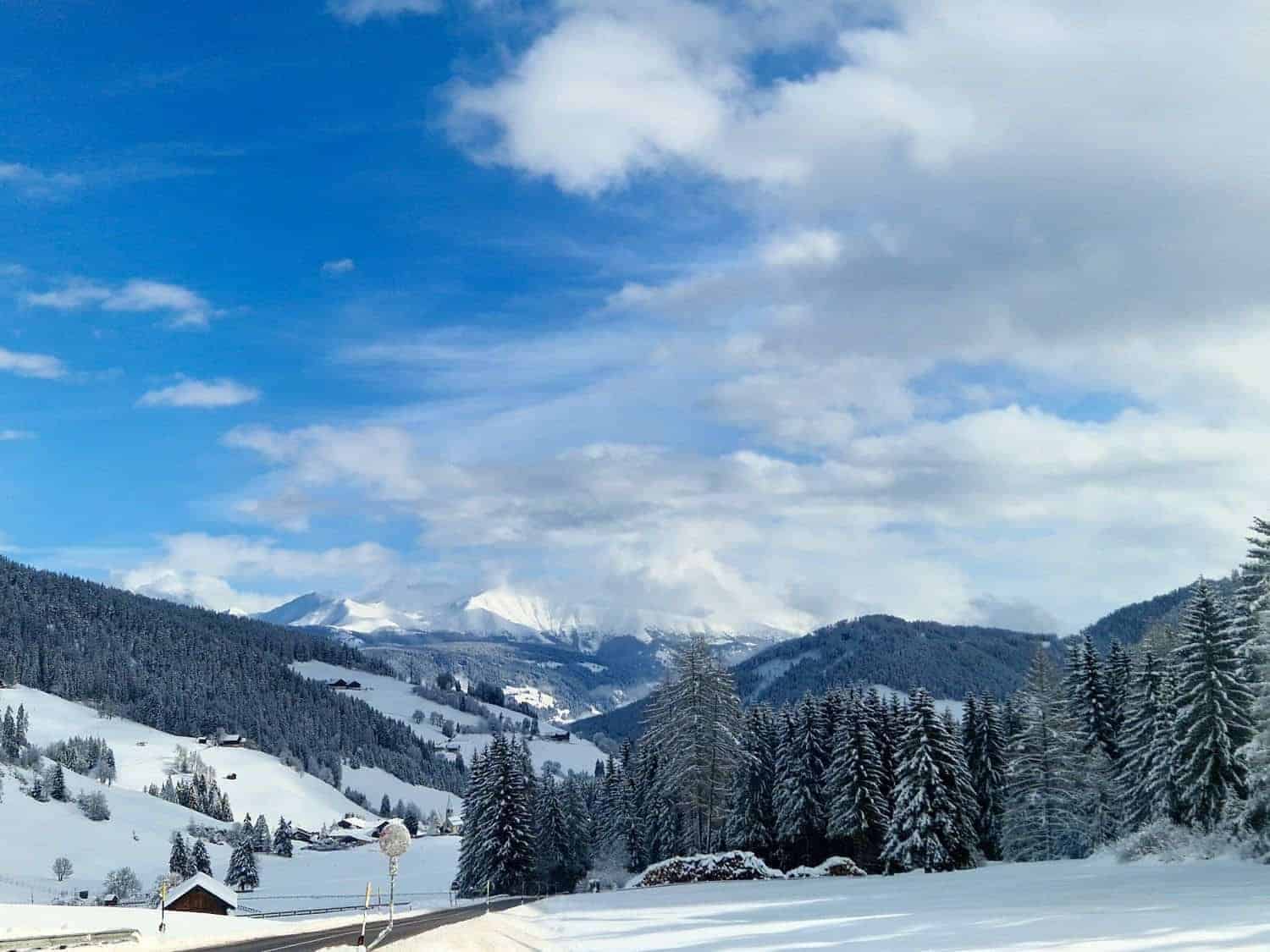 A scenic winter landscape showing snow-covered hills and pine trees under a blue, partly cloudy sky. Majestic mountains are visible in the distance, and a small wooden cabin sits near the foreground.