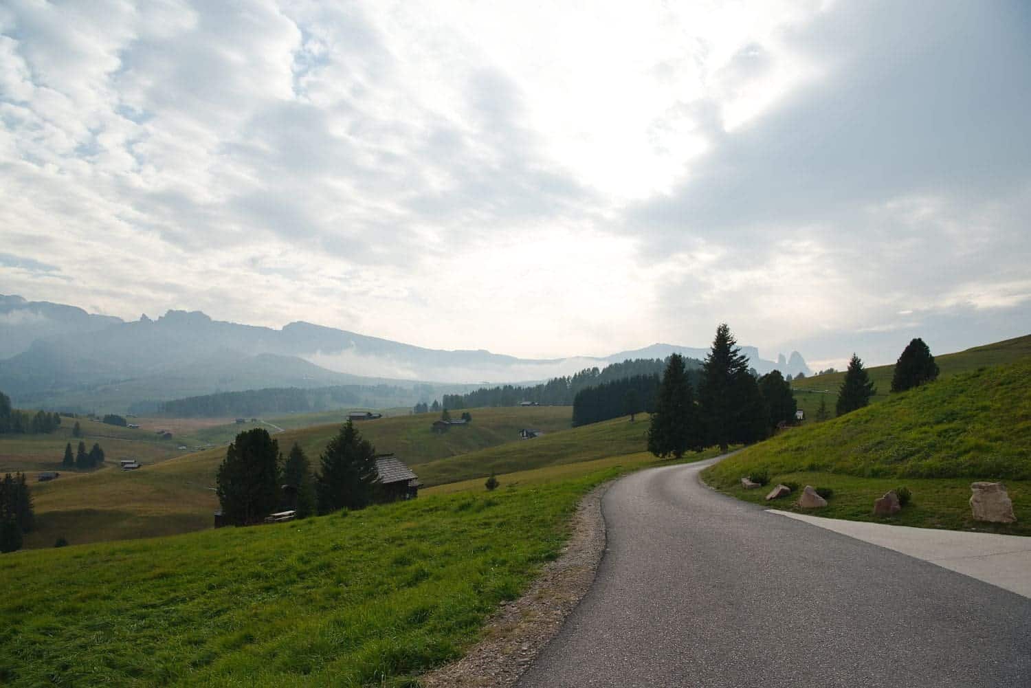 A winding road curves through a lush green landscape with scattered trees and small cabins. In the distance, misty mountains rise under a partly cloudy sky, creating a serene and picturesque scene.
