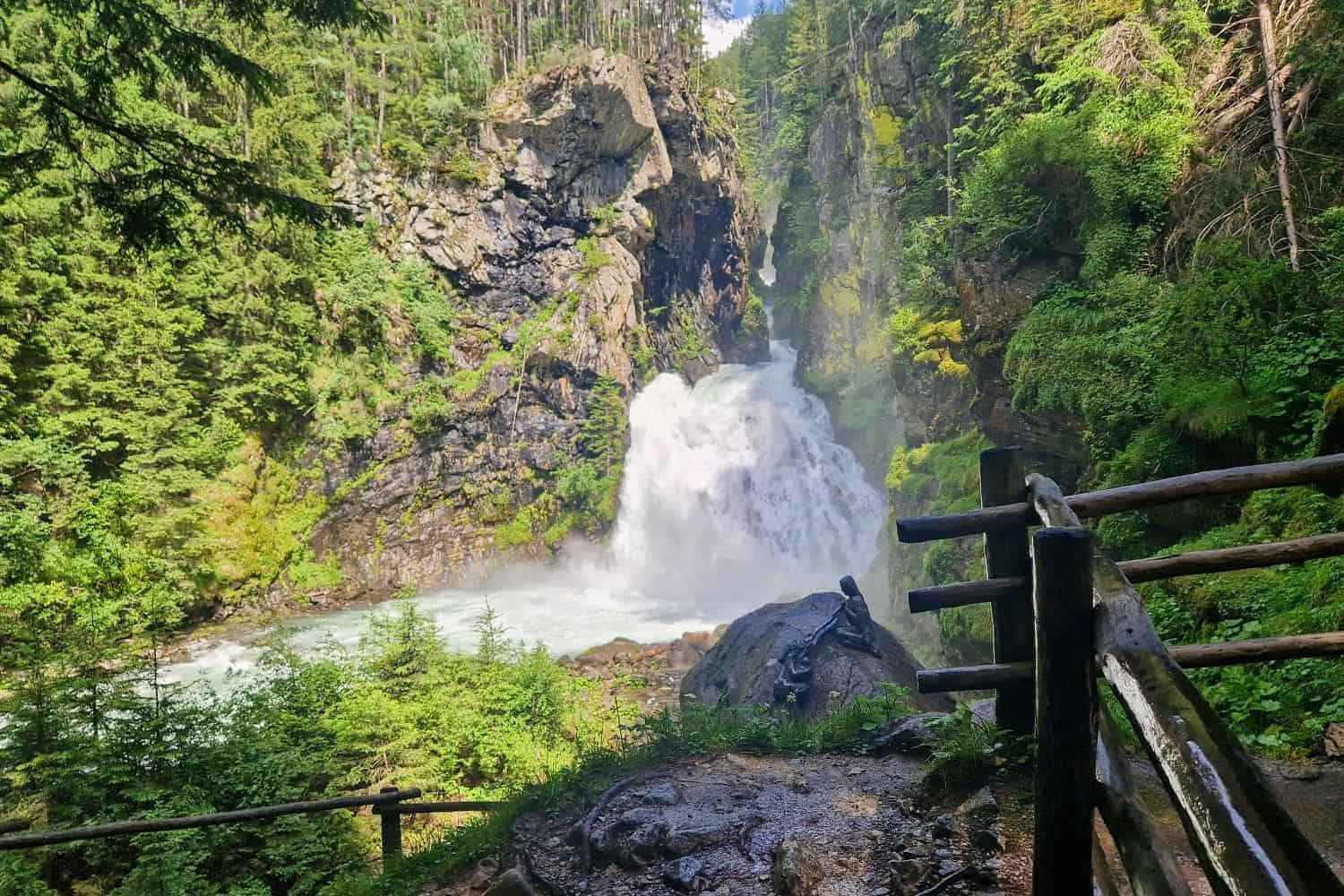 A scenic view of a waterfall cascading down a rocky cliff, surrounded by lush greenery and trees. A wooden fence lines a pathway in the foreground, leading to the waterfall. Sunlight filters through the trees, illuminating the scene.