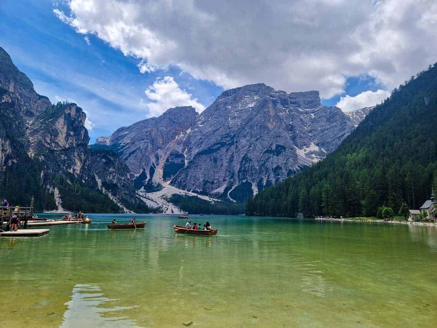 A serene lake with clear turquoise water surrounded by lush green forests and towering mountains under a partly cloudy sky. People in small boats paddle on the water, and a dock is visible on the left side.