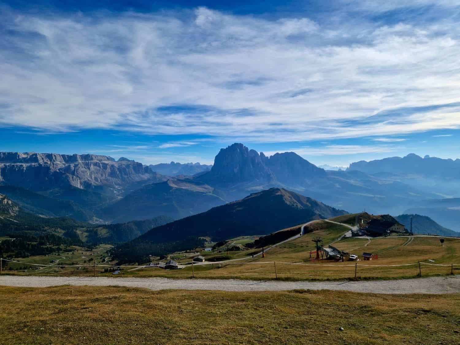 Scenic landscape of rolling hills and valleys with a rugged mountain range in the distance under a cloudy blue sky. The foreground features grassy fields and scattered paths, creating a tranquil, expansive view.