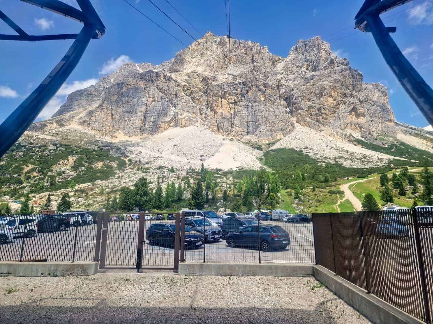 A rugged mountain under a blue sky towers over a parking area filled with cars. Green foliage covers parts of the landscape below the rocky cliffs. The foreground features a metal fence and structural components.
