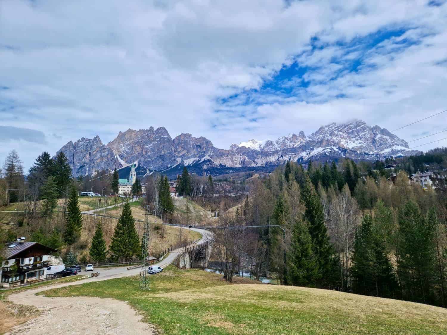 A scenic landscape features a winding road leading through a hilly area with sparse trees. In the background, rugged snow-capped mountains rise under a partly cloudy sky. A few houses and a small church are nestled among the trees.