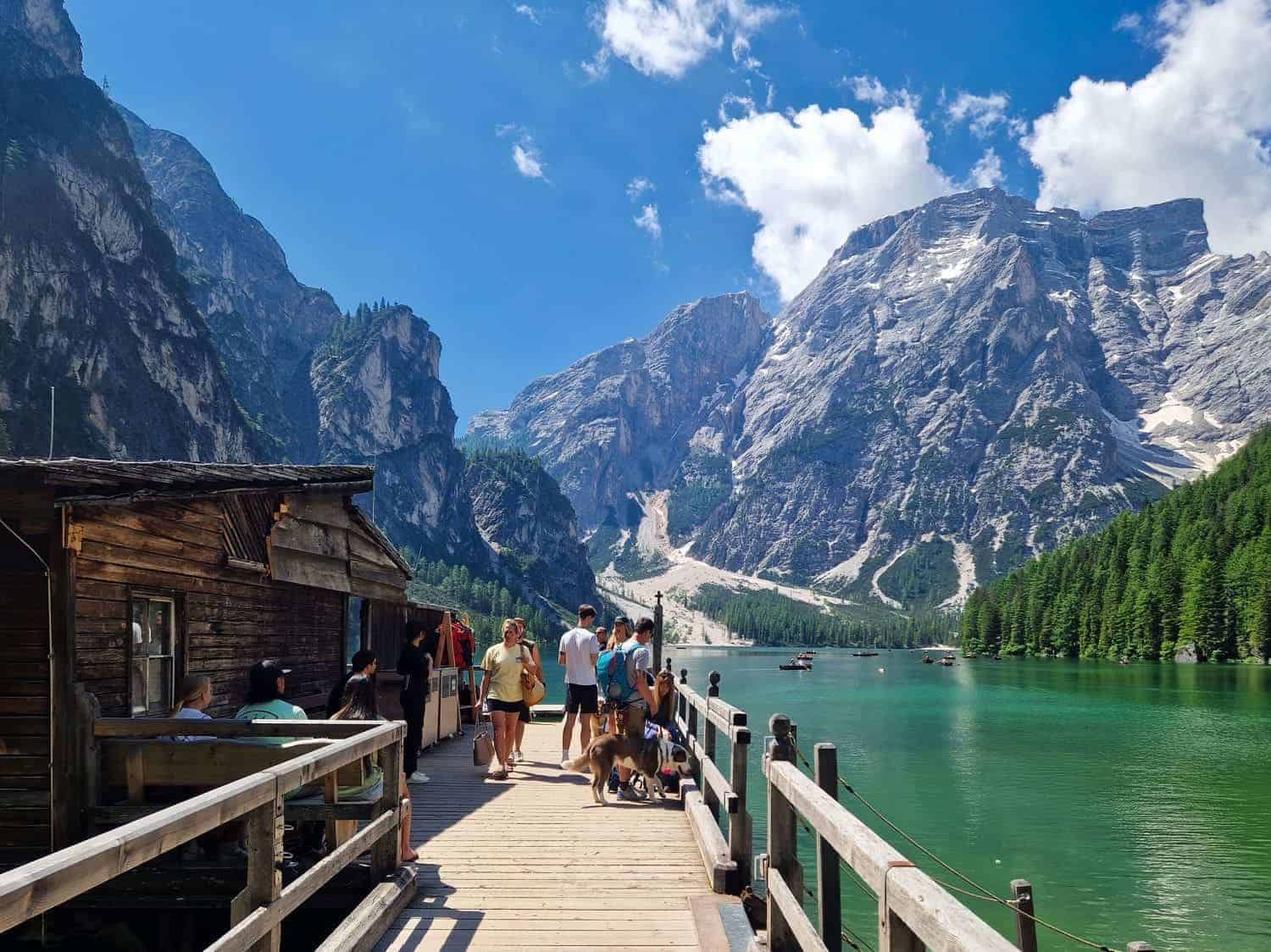 A mountain lake with turquoise water and wooded shores is surrounded by towering peaks. People walk on a wooden dock next to a rustic cabin. The scene is bright and sunny, with a few clouds in the blue sky.