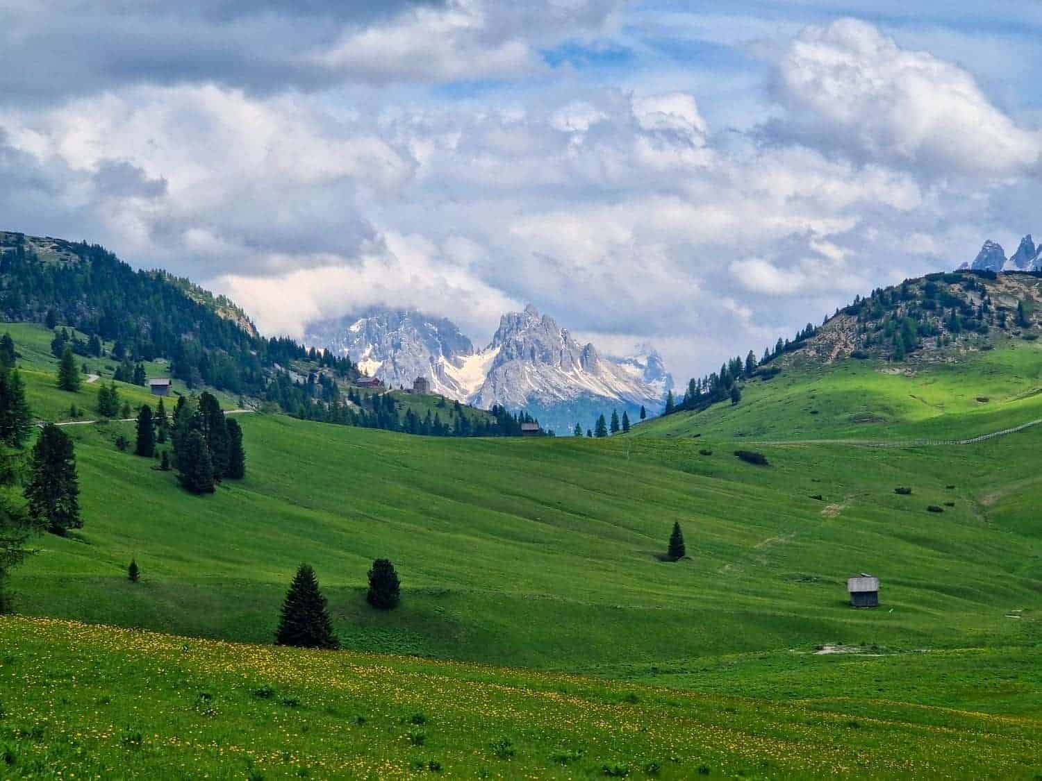 A scenic landscape featuring a lush green valley with scattered trees and a few huts. Rolling hills lead to distant, snow-capped mountains under a partly cloudy sky, creating a serene and picturesque view.