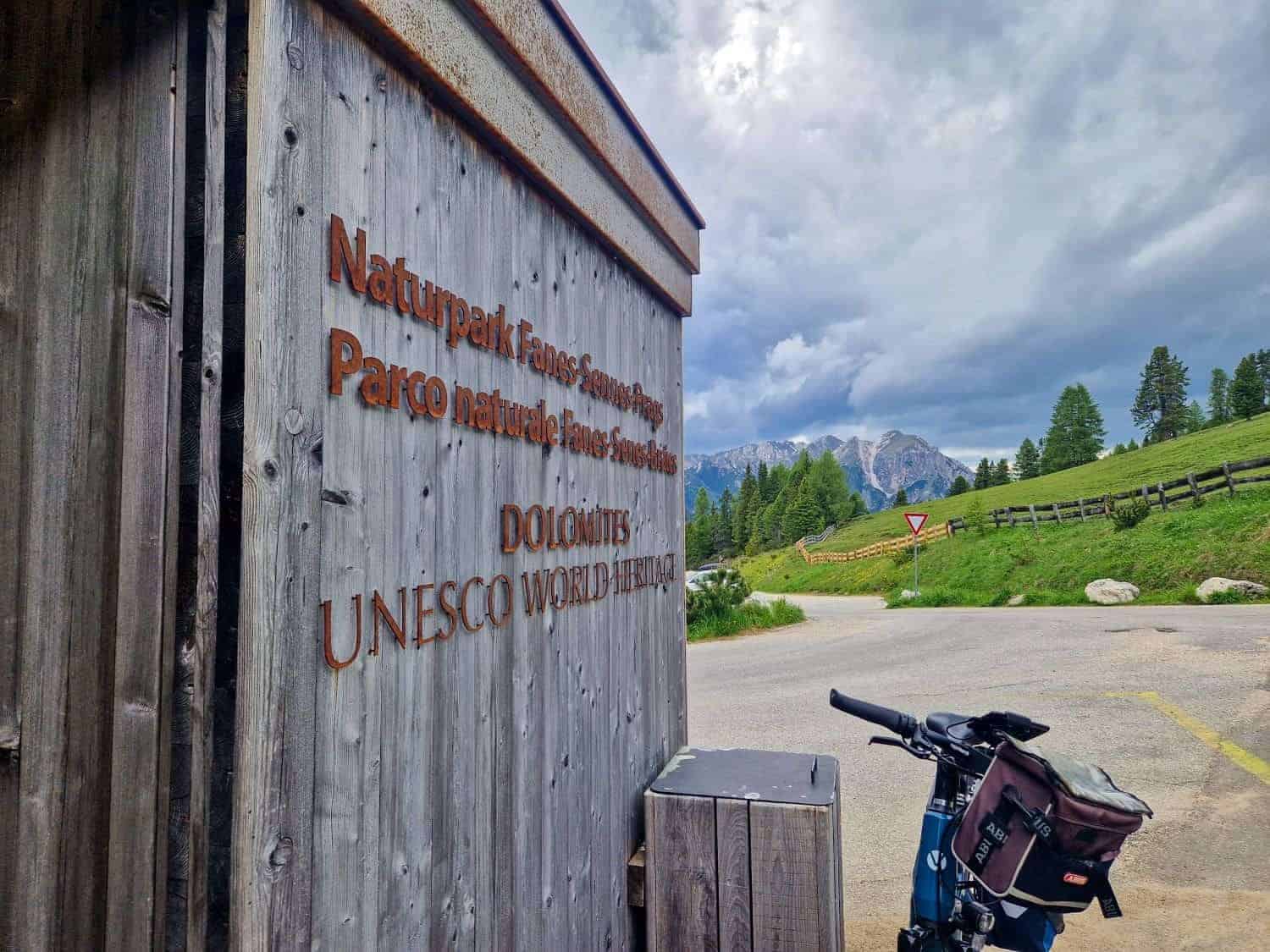 Wooden sign for "Naturpark Fanes-Sennes-Prags" in the Dolomites, a UNESCO World Heritage site. A bicycle is parked nearby. The view includes a road, a grassy area, and a distant mountain range under a cloudy sky.