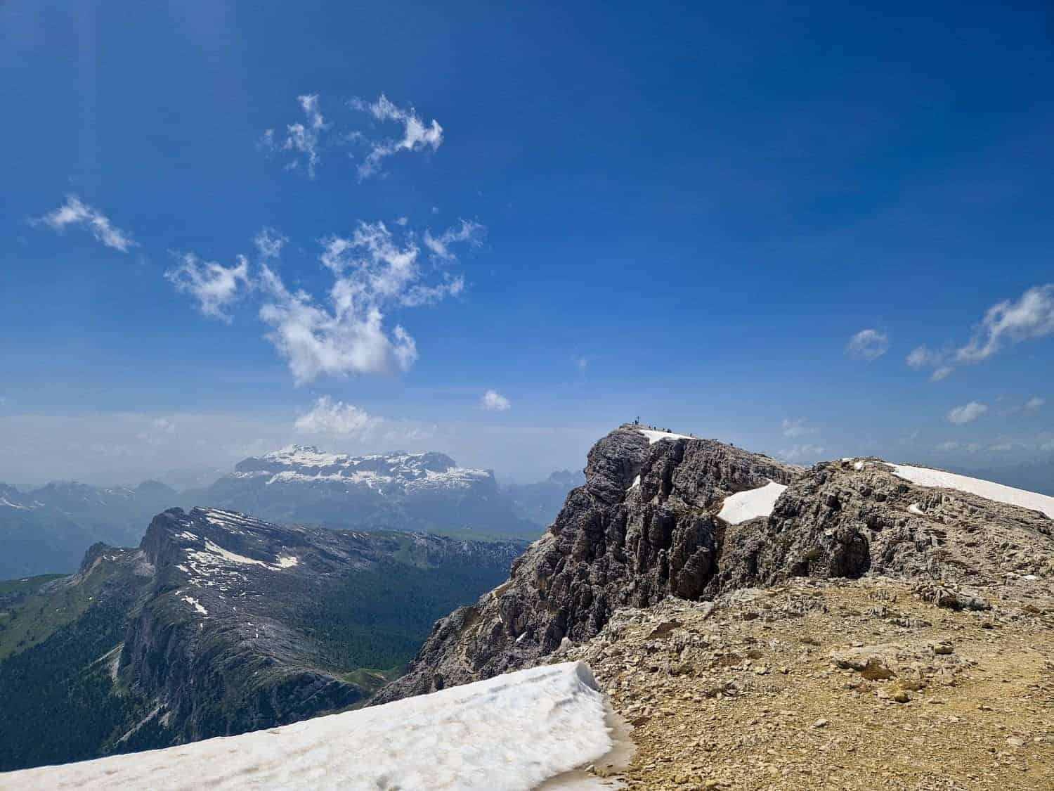 A breathtaking view of a rocky mountain landscape under a bright blue sky with scattered clouds. Snow patches are visible on the ground and mountain peaks, with lush green valleys in the background.