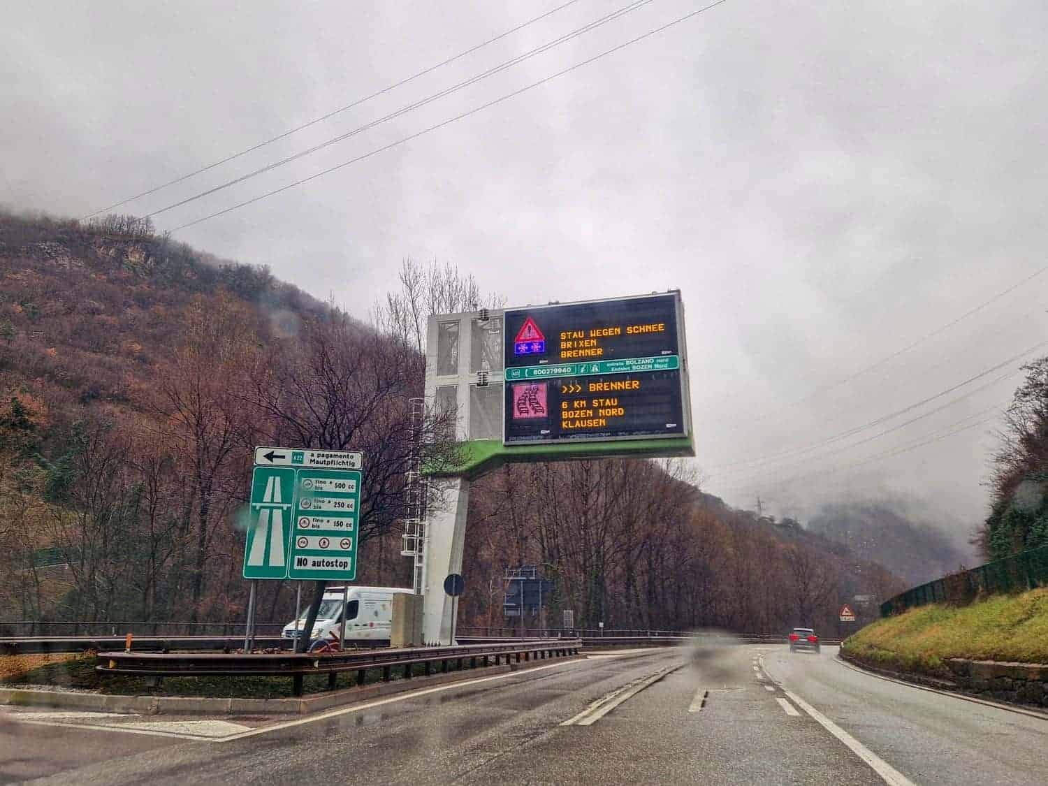 Highway scene on a rainy day with traffic signs in multiple languages. Surrounding landscape features misty hills and bare trees. Vehicles are visible on the wet road. The sky is overcast with clouds.