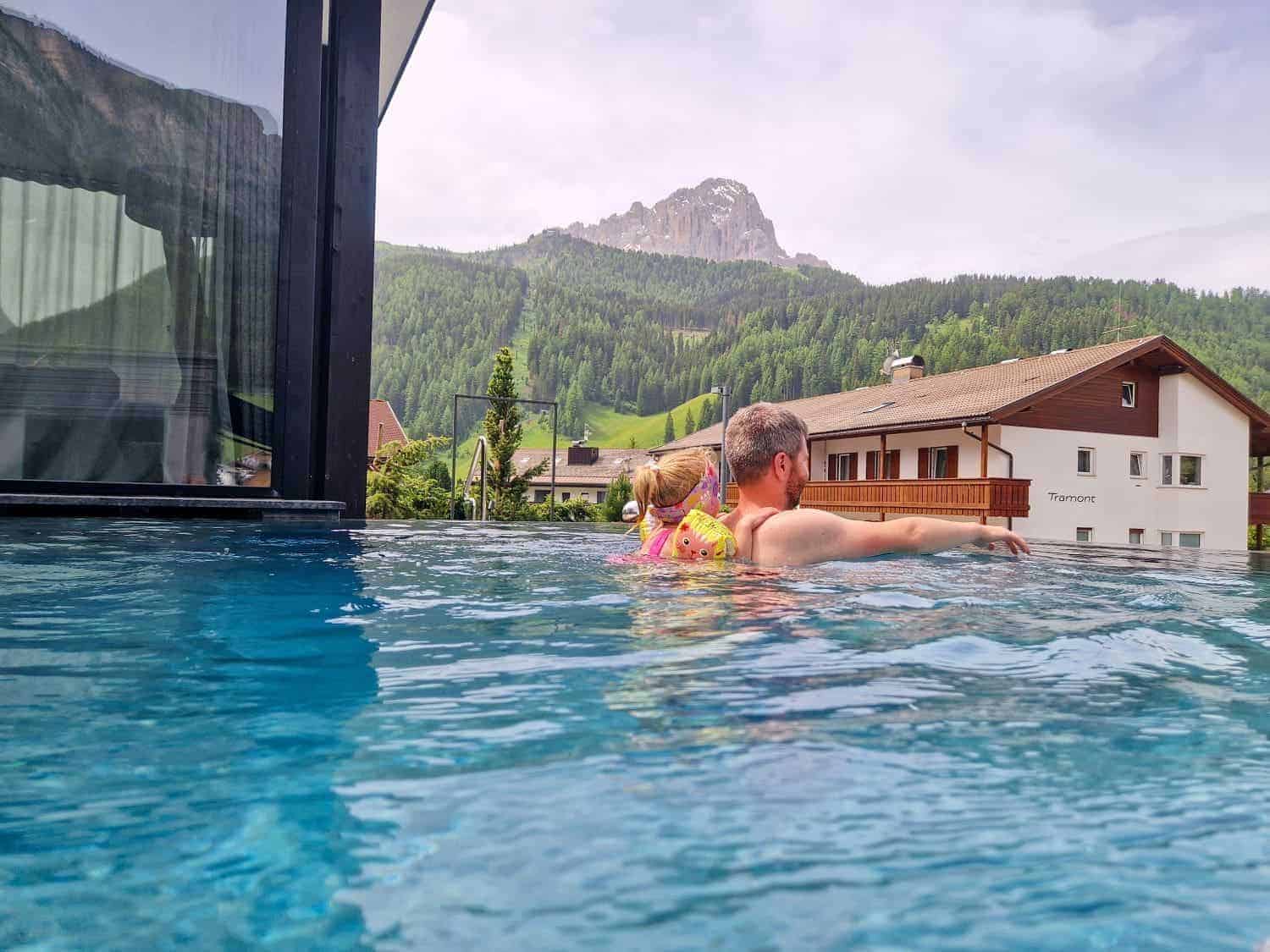 A man and a child in a pink swimsuit enjoy a modern outdoor pool with patio furniture visible. In the background, there are wooden houses, lush green trees, and a mountain under a cloudy sky.