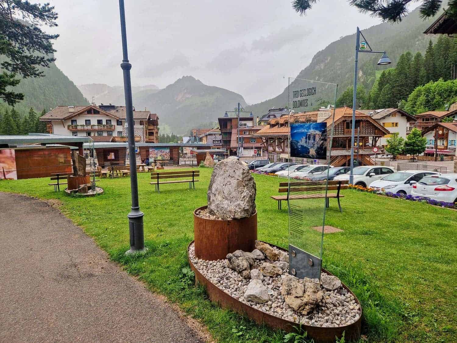 Scenic mountain village with green grass, benches, and a rock display in the foreground. Rustic buildings line the street with parked cars. Mountains and trees form the backdrop under a cloudy sky.