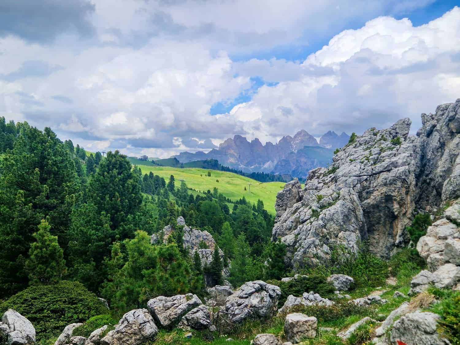 Scenic view of the Dolomites with a mix of rocky terrain and lush green hills. Cloudy sky above creates dramatic lighting, and distant mountains are partly visible in the background. Large rocks and pine trees frame the landscape.