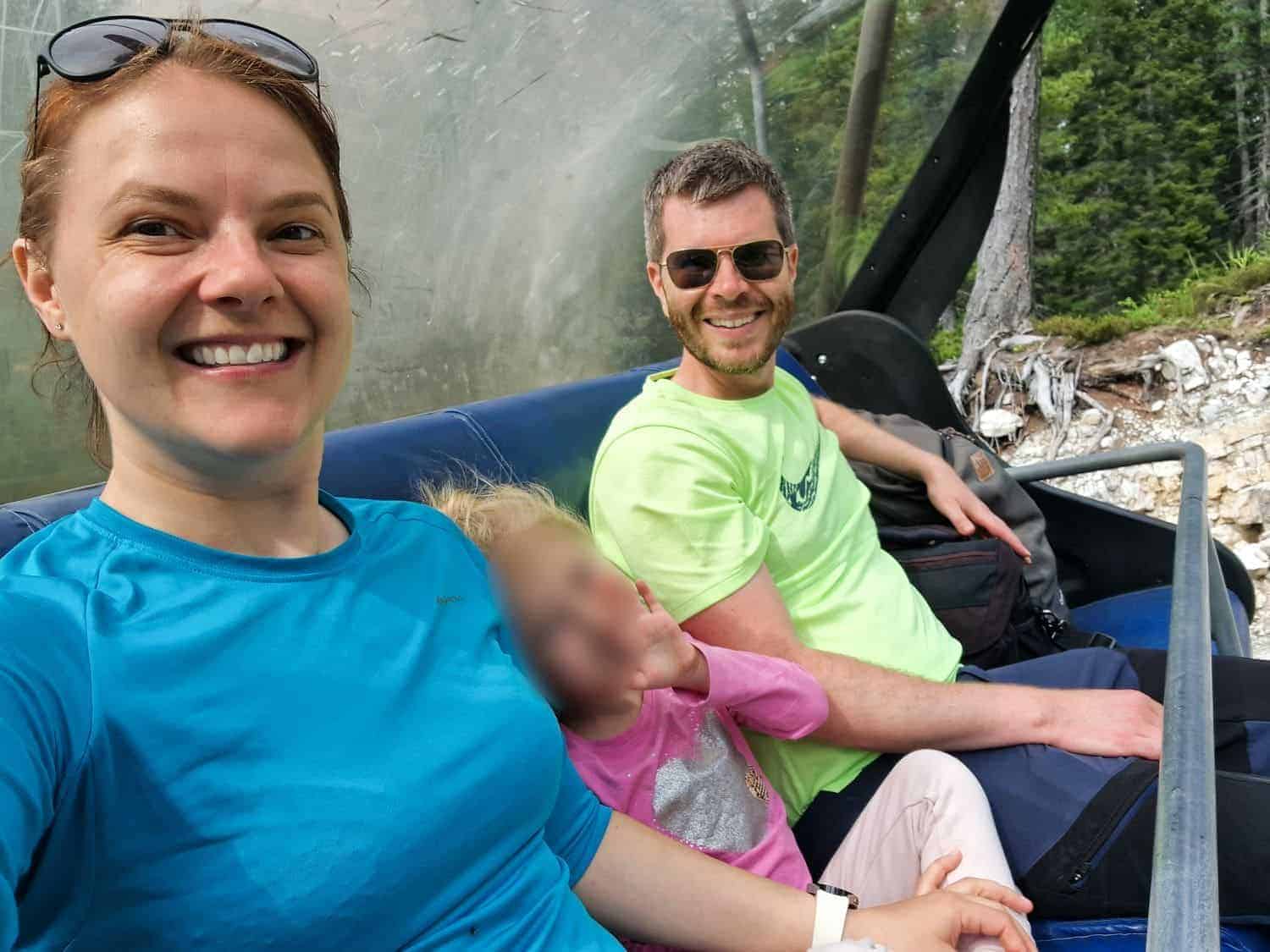 A woman and a man sit on a chairlift, smiling at the camera. The woman wears a blue shirt and sunglasses on her head; the man wears a light green shirt and sunglasses. A young child in pink is between them, face blurred. Trees are in the background.