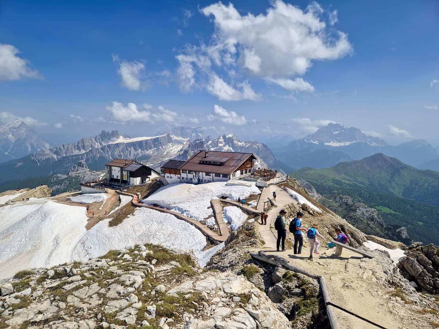 A scenic view of a mountainous landscape with snow patches. A group of hikers on a trail leads to a cabin on the ridge. Majestic mountain peaks and a partly cloudy sky form the backdrop.