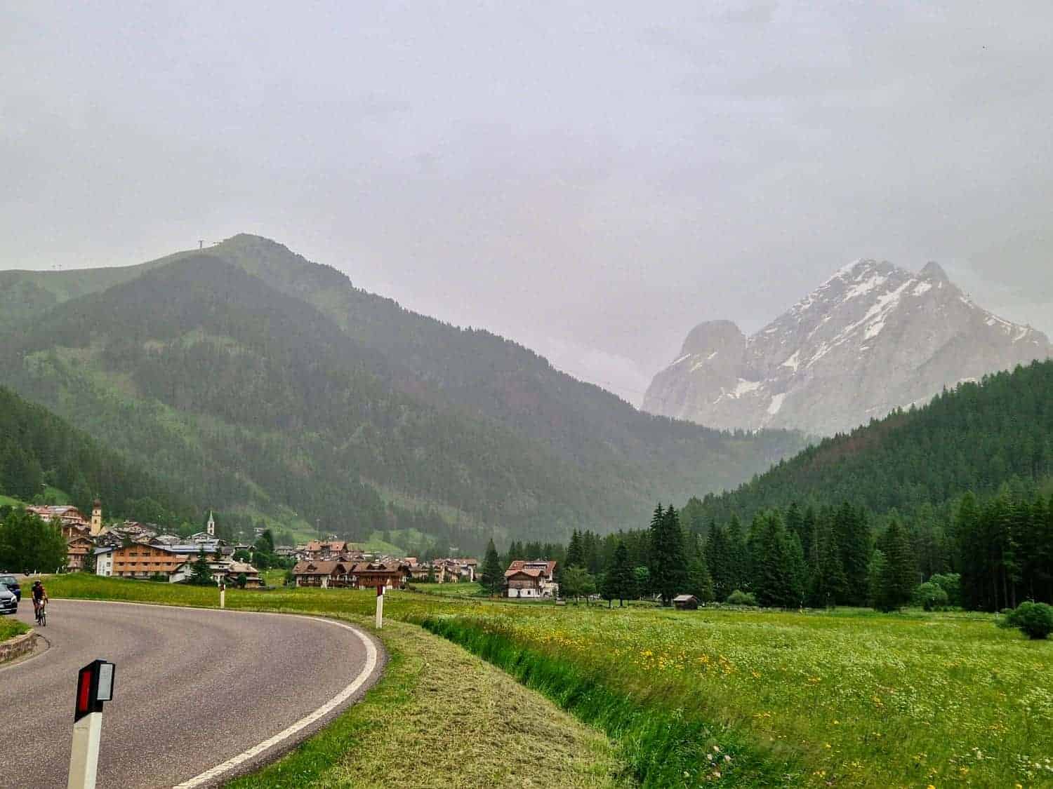 Winding road leads to a quaint village nestled in a lush green valley. Snow-capped mountains rise in the background under a cloudy sky. A cyclist rides along the road, surrounded by fields and forests.
