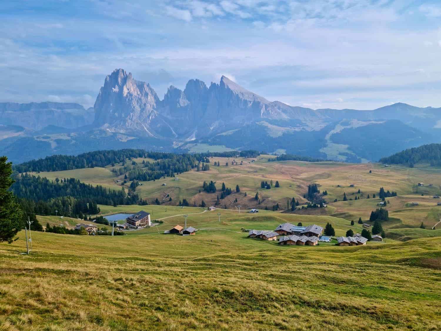 Mountain landscape featuring grassy meadows, scattered farmhouses, and a cluster of buildings. Tall mountains with jagged peaks rise in the background under a blue sky with soft clouds. Trees are dotted across the rolling landscape.