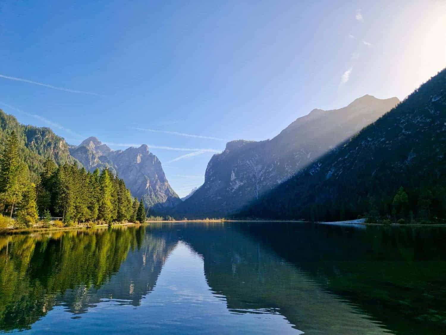 A serene lake reflecting surrounding pine forest and steep mountains under a clear blue sky. Sunlight casts a shadow on part of the mountain, creating a tranquil and picturesque landscape.