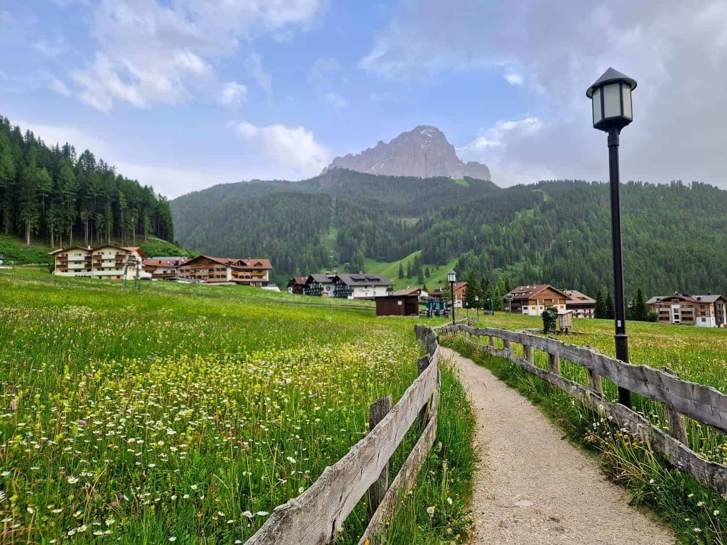 A scenic view of a pathway bordered by a wooden fence, leading through a green meadow full of wildflowers. In the background, there are houses near a forested area, with a mountain rising under a partly cloudy sky. A lamp post stands on the right.