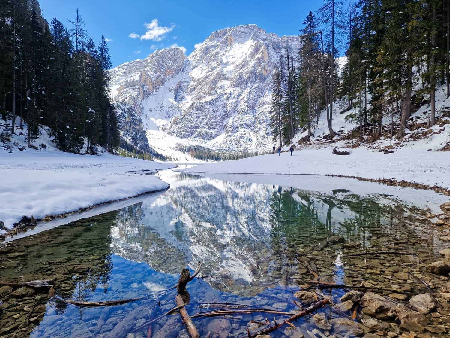 A clear mountain lake reflects snow-covered peaks and surrounding evergreen trees. The foreground shows stones and branches in shallow water. A few people are walking along the snowy path by the lake under a bright blue sky.