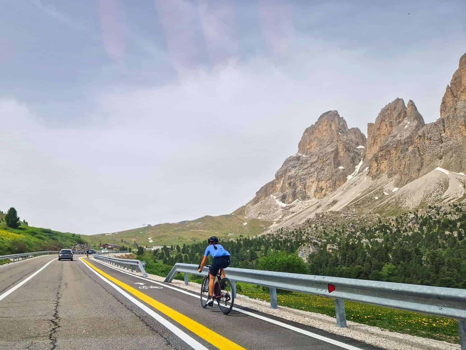 A cyclist wearing a blue shirt rides along a road surrounded by mountainous terrain. The road features distinct bike and car lanes. In the background, large rocky peaks rise under a partly cloudy sky.