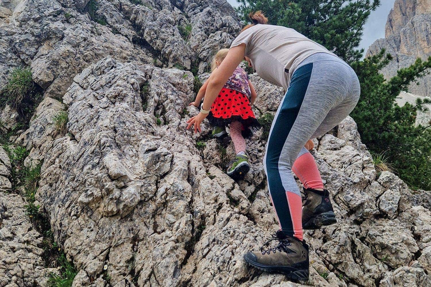 A woman and a child in a red polka dot dress climb a rocky hillside. The woman wears a white shirt, leggings, and hiking boots. The landscape includes rugged rocks and a tree in the background.