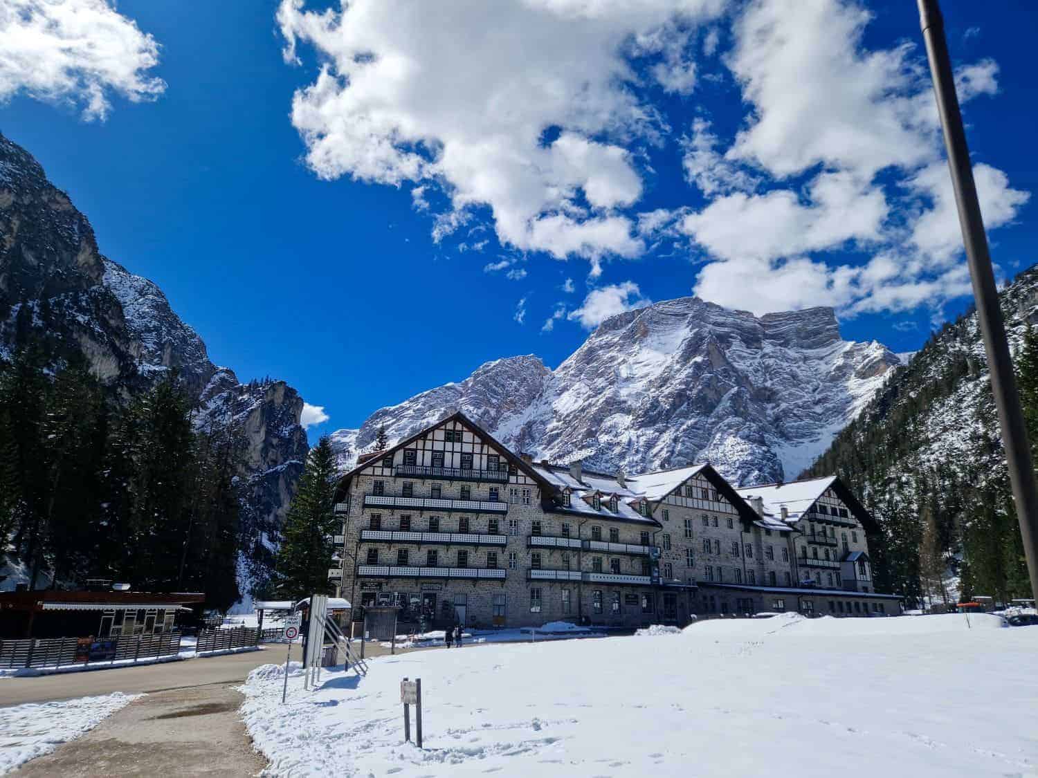A large, alpine-style hotel stands amidst a snowy landscape with towering mountains in the background. The sky is bright blue with scattered fluffy clouds. Pine trees flank the building, adding to the picturesque, serene setting.
