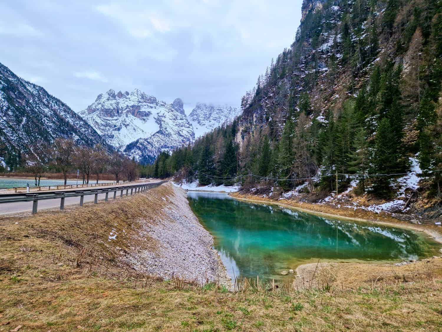 A serene turquoise pond sits beside a mountain road with a wooden guardrail. Snow-dusted trees line the surrounding rugged terrain, and snow-covered peaks rise in the background under a cloudy sky.