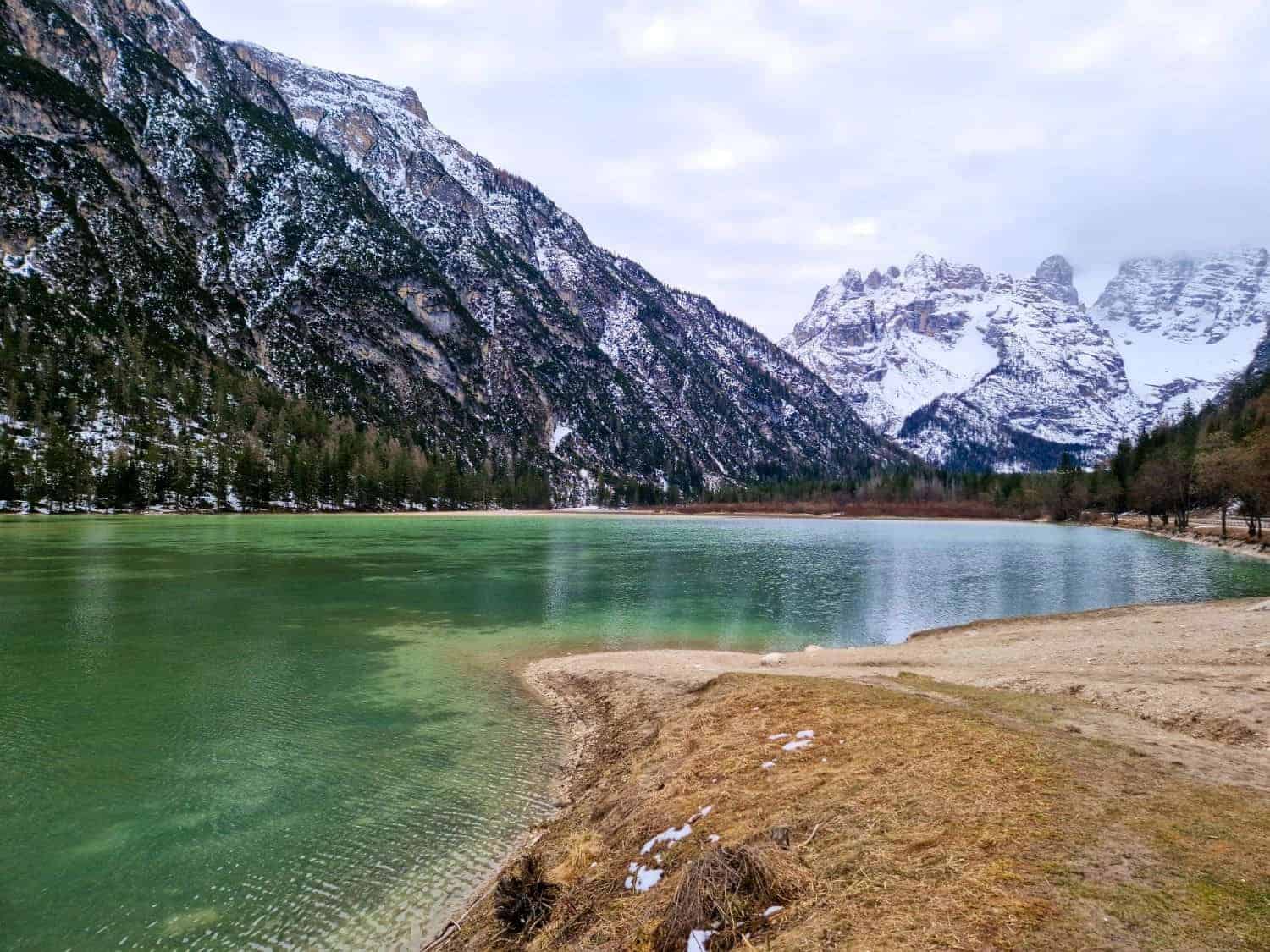 A serene mountain lake with green waters is surrounded by snow-dusted mountains. A grassy, sandy shore curves along the lake's edge, and tall trees line the opposite bank under a cloudy sky.