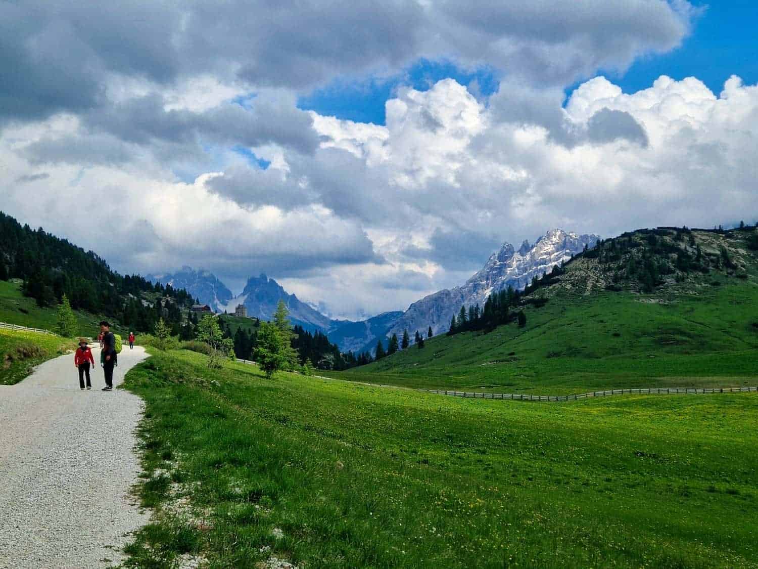 A group of hikers walk along a gravel path through a lush green meadow, surrounded by hills and scattered trees. Snow-capped mountains and a dramatic, cloudy sky form the backdrop.