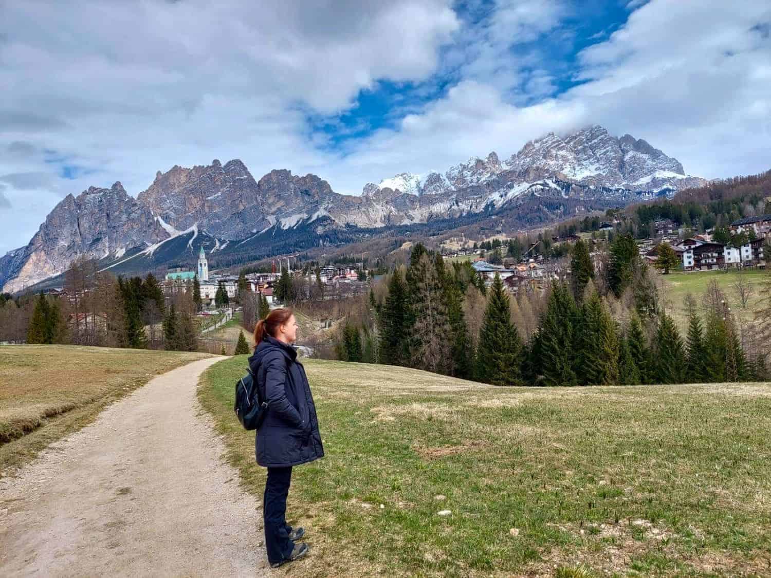 A person in a dark coat and backpack stands on a dirt path, gazing at a mountainous landscape dotted with trees and houses. Snow-capped peaks rise in the distance under a partly cloudy sky.