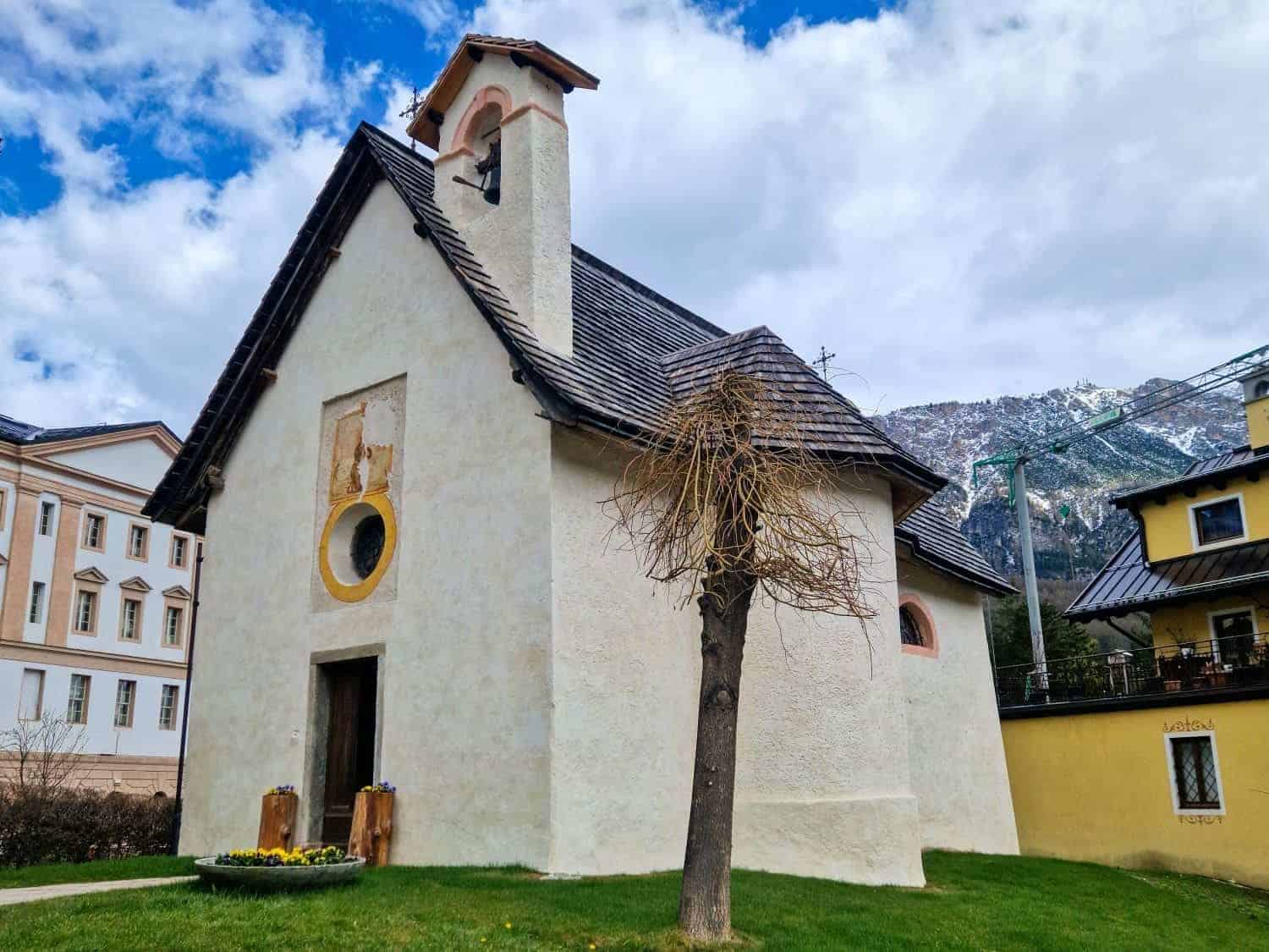Small white chapel with a sloped roof and bell tower, surrounded by a grassy area. A leafless tree stands in front. Snow-capped mountains are visible in the background under a partly cloudy sky.