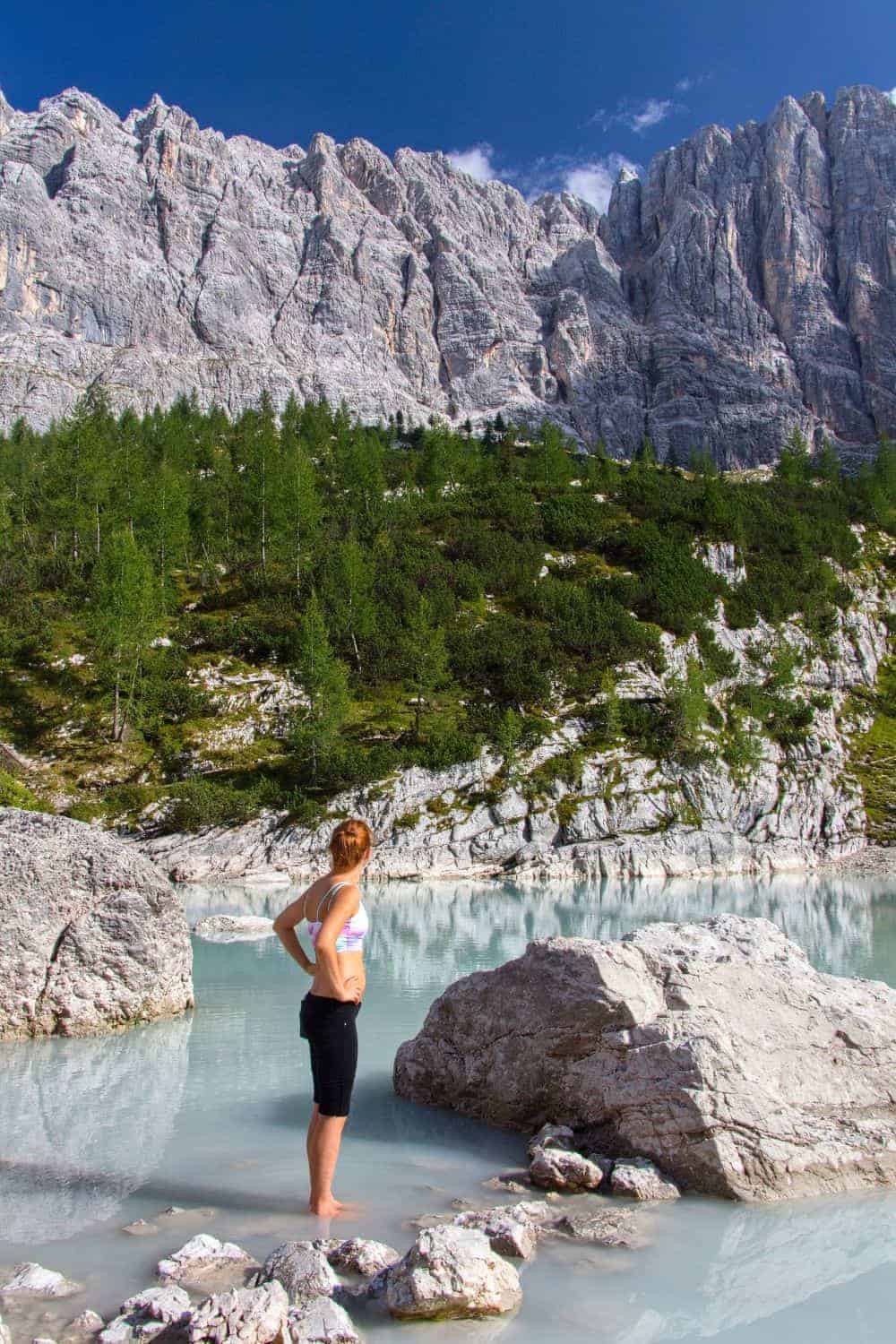 A woman stands on rocks at the edge of a serene, turquoise lake. She gazes towards towering, rugged mountains under a bright blue sky. The landscape is lush with green trees. She is wearing activewear and has her hands on her hips.