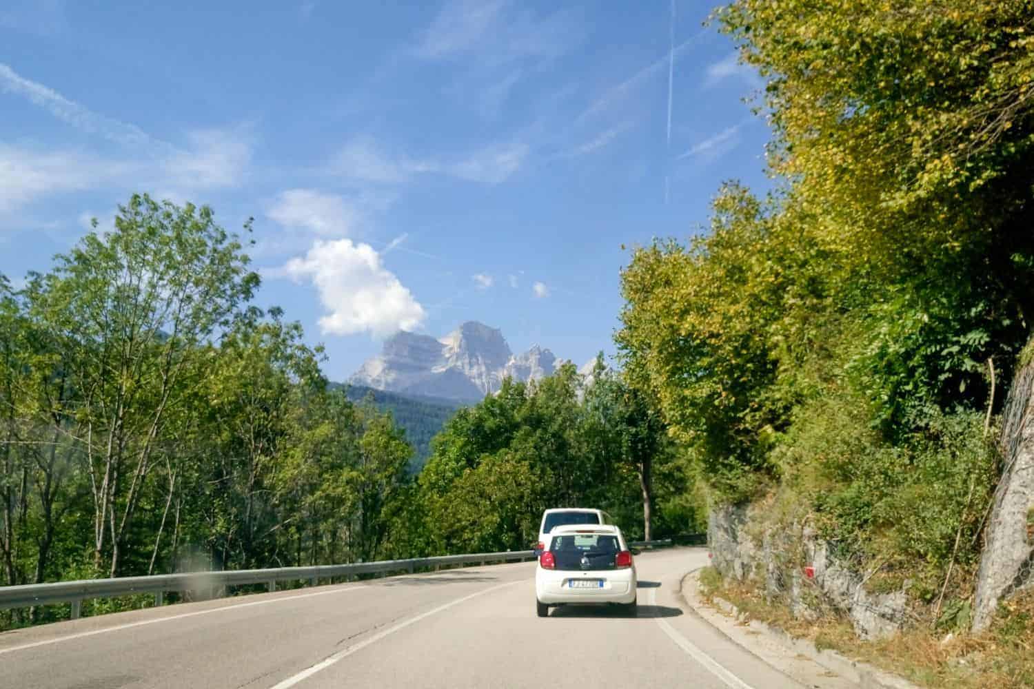 A white car drives on a winding road bordered by green trees. In the background, a mountain with a partly cloudy sky is visible. The road curves gently to the left, and the scene is under a bright, clear sky.