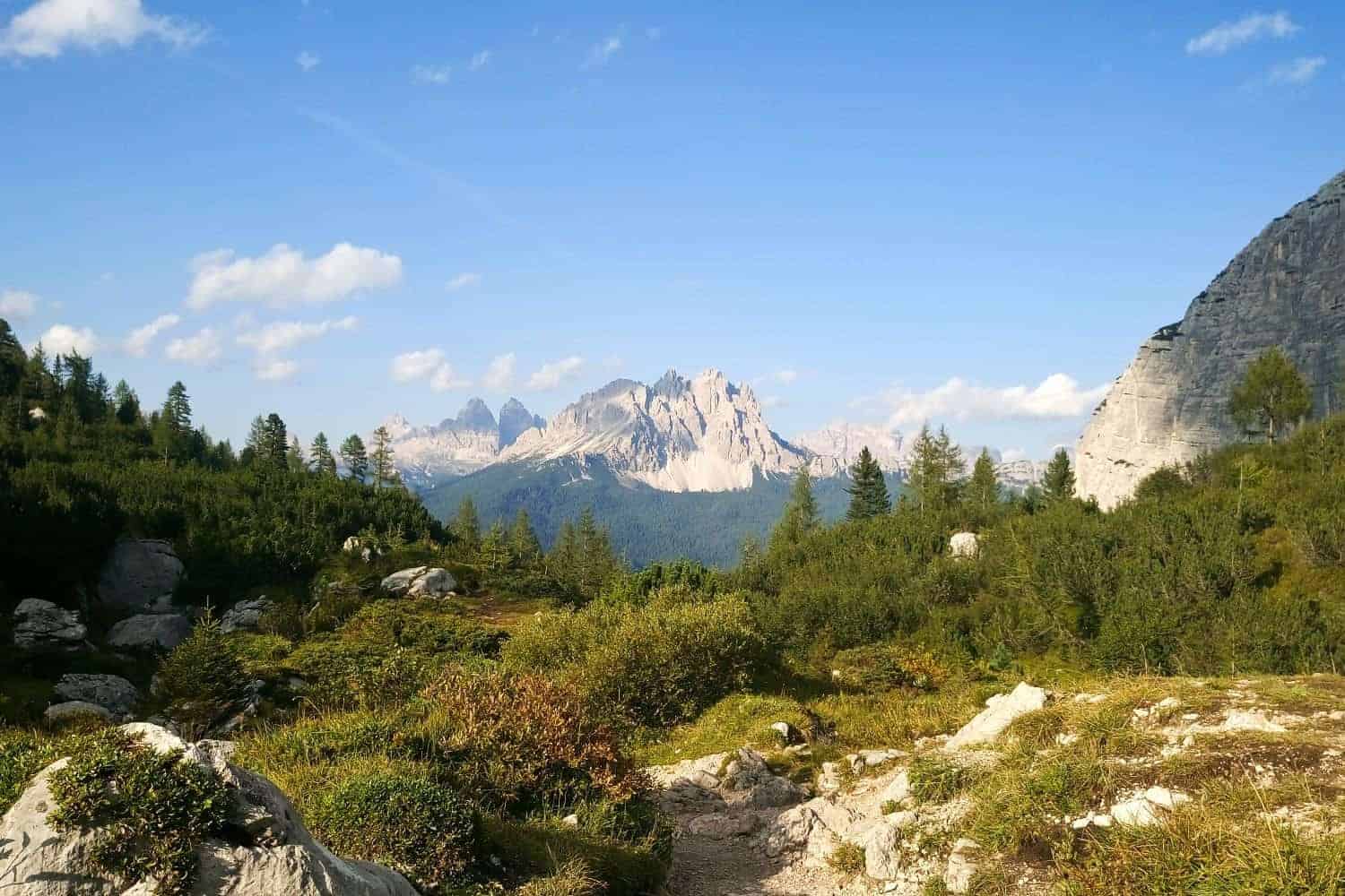 Scenic view of a mountainous landscape, featuring rugged peaks in the distance under a blue sky with scattered clouds. The foreground includes greenery, rocky terrain, and a pathway leading towards the horizon.