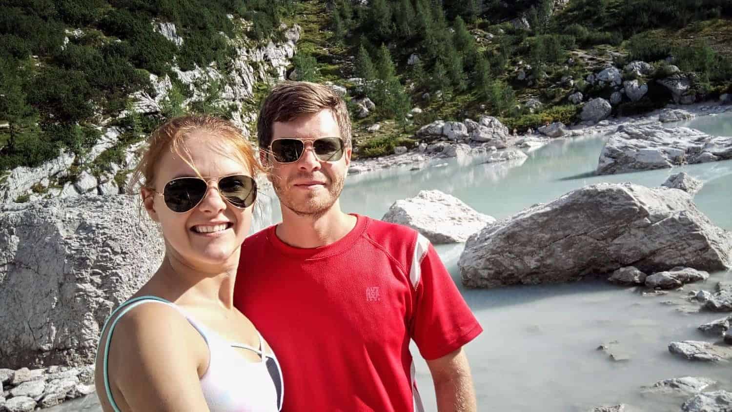 A smiling couple wearing sunglasses poses for a selfie in front of a rocky landscape with a serene, milky blue lake. Green vegetation covers the slopes in the background.
