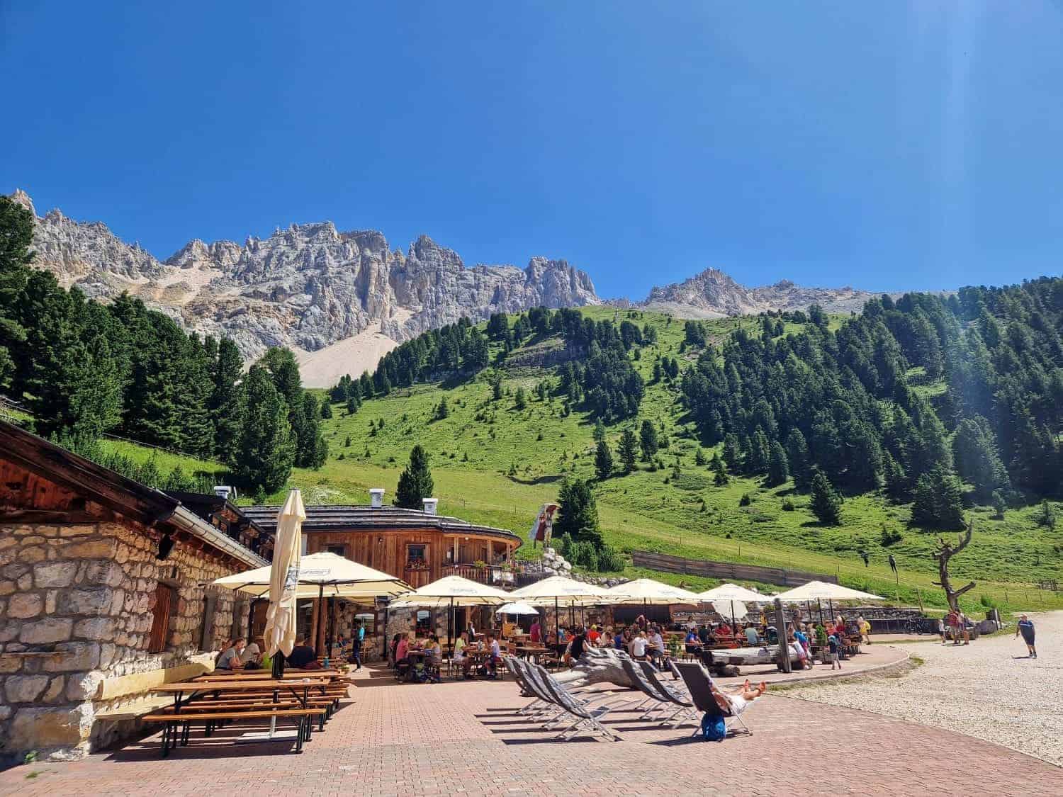 Outdoor seating area with umbrellas and lounge chairs on a sunny day, surrounded by green hills and pine trees. A rocky mountain forms the backdrop under a clear blue sky. People are relaxing and enjoying the scenery.