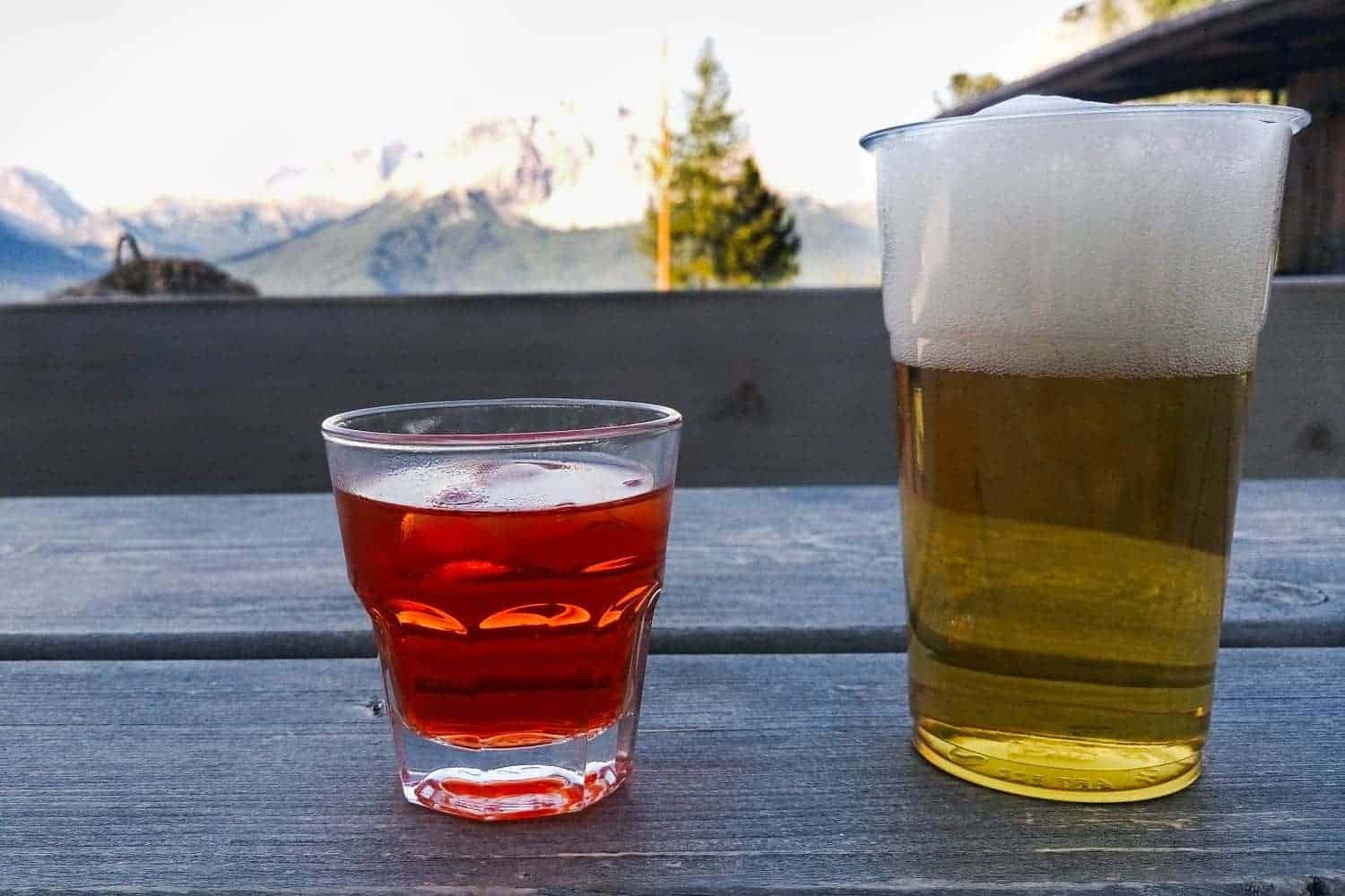 A glass of orange-colored aperitif with ice and a glass of frothy beer sit on a wooden table. In the background, there are blurry views of mountains and trees under a clear sky.
