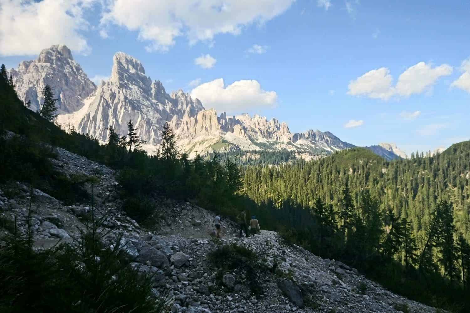 A scenic view of a mountainous landscape with rugged peaks under a blue sky. Pine trees cover the slopes, and two hikers walk along a rocky trail in the foreground. Fluffy clouds dot the sky, adding a serene touch to the panorama.
