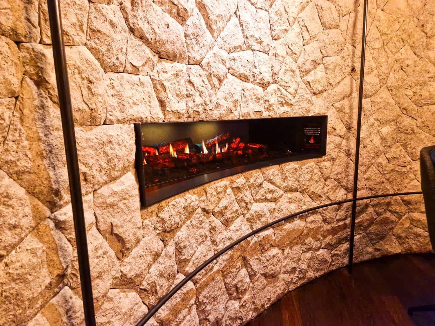 Modern electric fireplace set in a curved stone wall, displaying realistic glowing embers and faux logs. The surrounding stones are textured and light-colored, adding a rustic contrast to the sleek design of the fireplace.