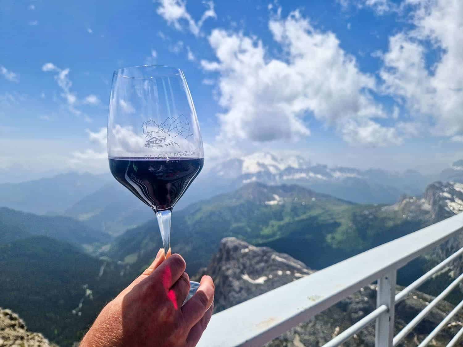 A person holds a glass of red wine against a backdrop of a mountainous landscape with lush green valleys and snow-capped peaks under a blue sky with scattered clouds. The glass features a mountain logo.