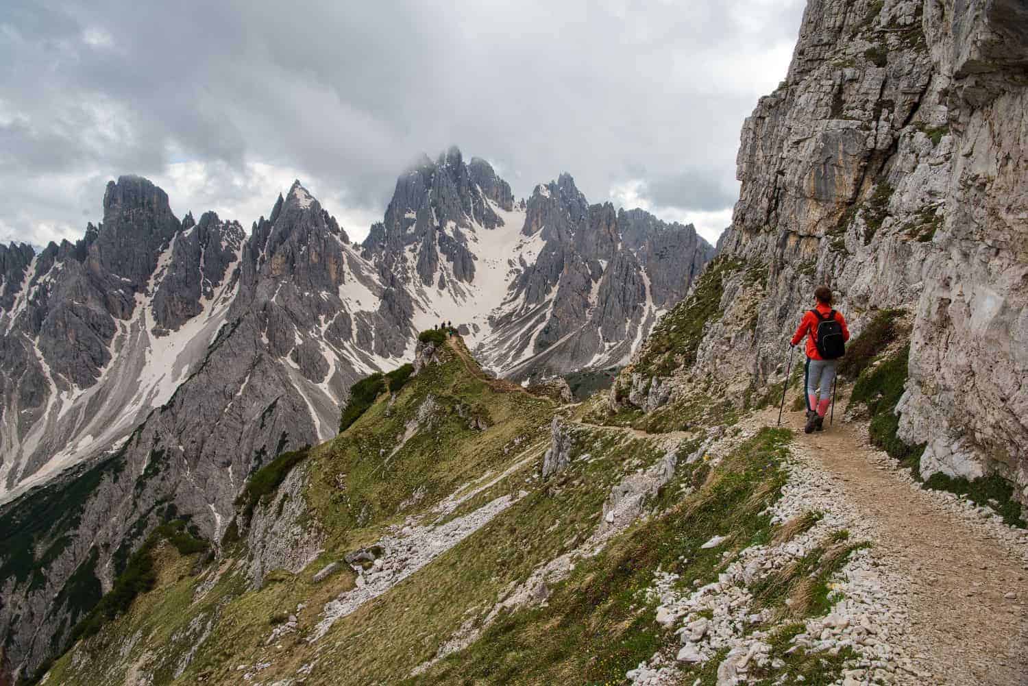 A hiker in a red jacket walks along a narrow mountain trail, surrounded by rugged rocky peaks partially covered in snow. The sky is cloudy, creating a dramatic backdrop for the towering mountains.