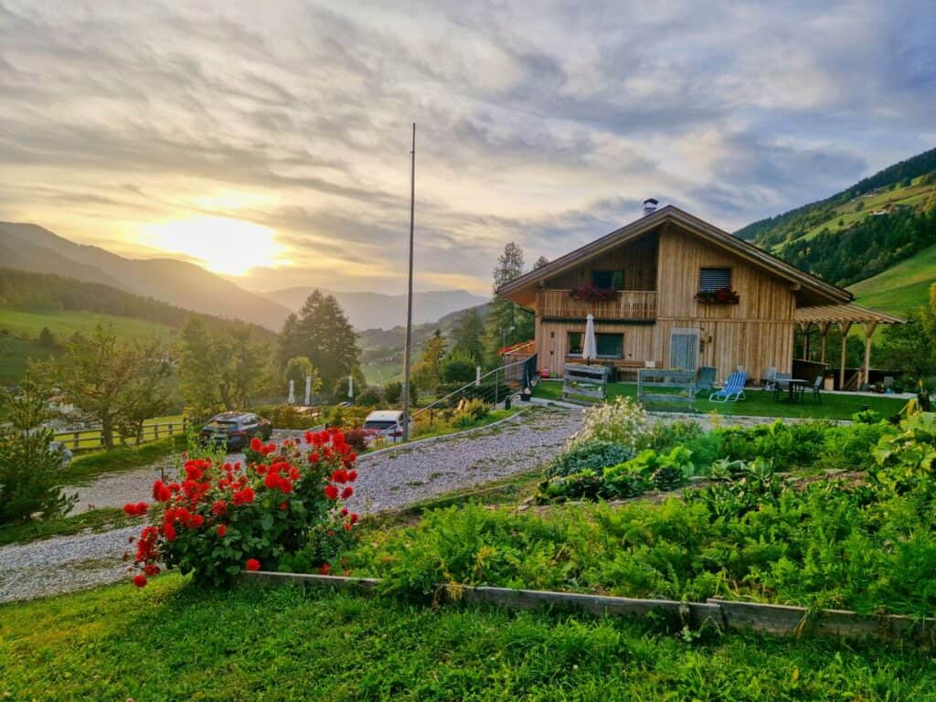 A wooden house sits on a green hillside garden with red flowers in the foreground, cars parked on a gravel driveway, and mountains in the background under a cloudy sunset sky.