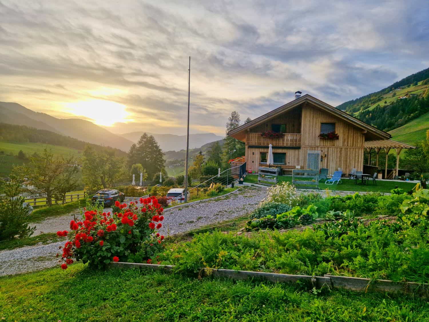 A wooden house sits on a green hillside garden with red flowers in the foreground, cars parked on a gravel driveway, and mountains in the background under a cloudy sunset sky.
