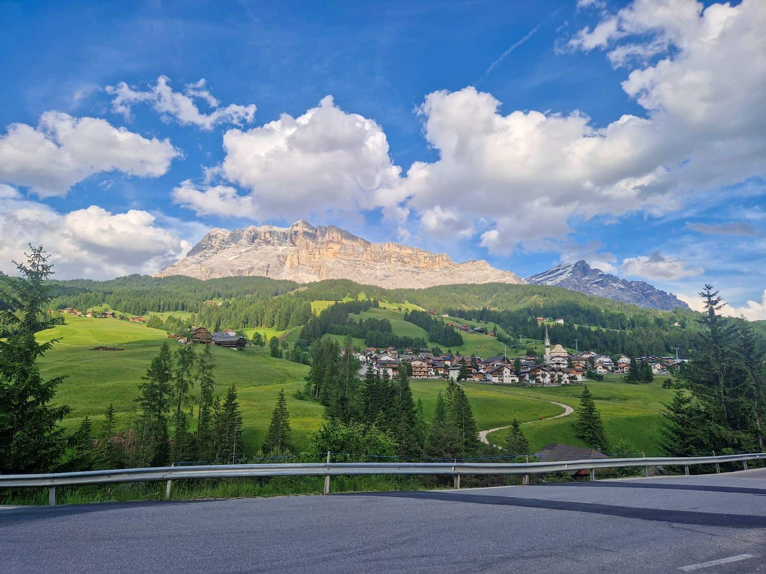 A scenic view of a small village nestled in green hills with pine trees, surrounded by mountains under a blue sky with fluffy white clouds. A road with a metal guardrail runs in the foreground.