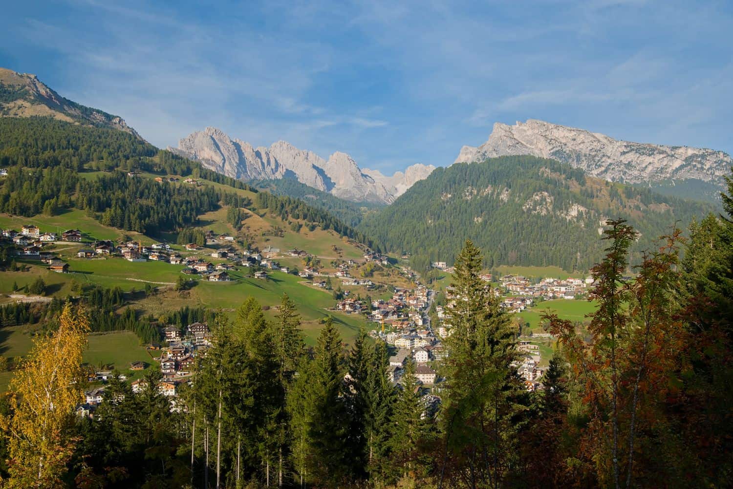 A scenic mountain village with scattered houses on green slopes, dense forests in the foreground, and rocky peaks rising under a blue sky in the background.