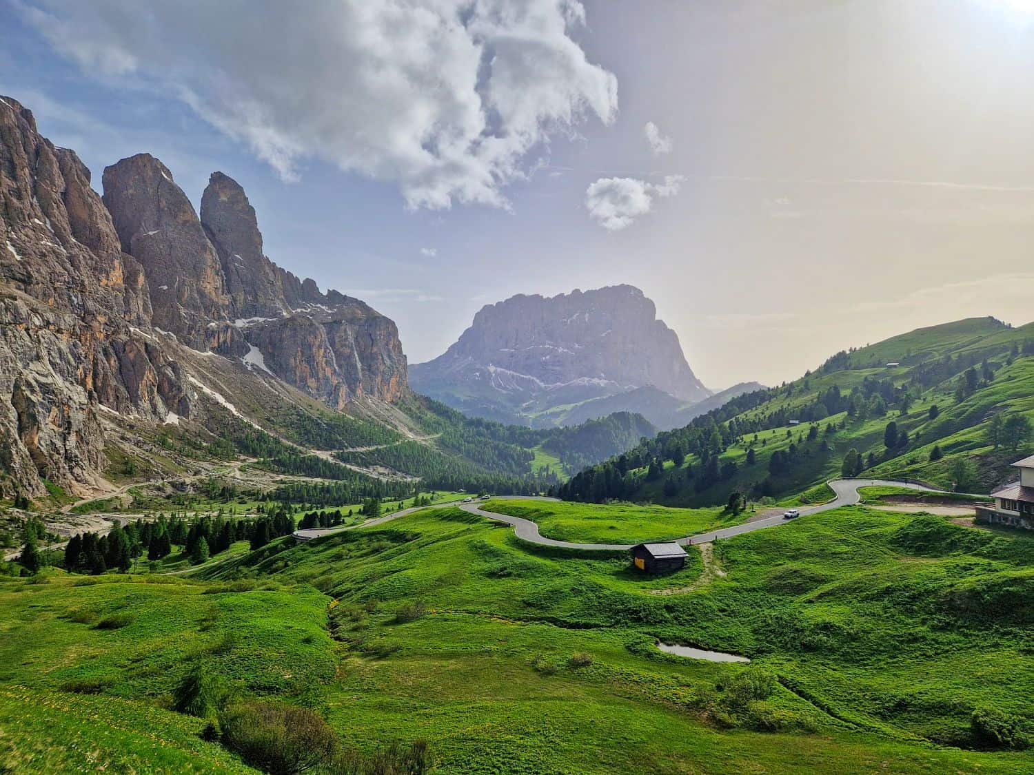A winding road passes through lush green hills, with rocky mountains rising in the background under a partly cloudy sky and sunlight streaming from the right. A small cabin sits beside the road in the picturesque landscape.