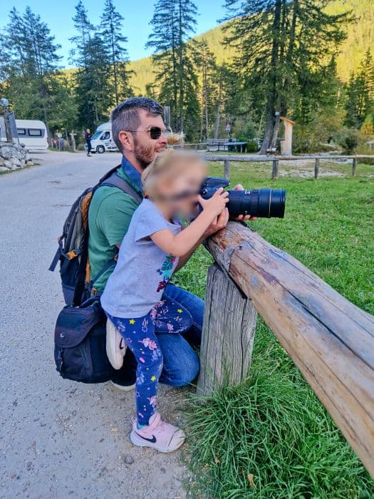 An adult kneels next to a child, helping them hold a large camera on a wooden fence in a park. Trees and a parked camper van are visible in the background under a sunny sky.