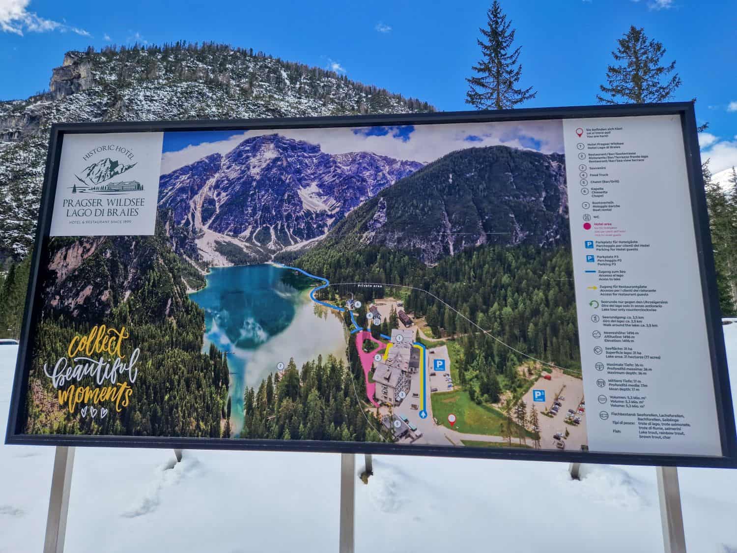 Large informational sign in a snowy landscape showing a map of Lago di Braies (Pragser Wildsee) with a photo of the lake, walking paths, buildings, legends, and the phrase “collect beautiful moments.” Pine trees and mountains are in the background.