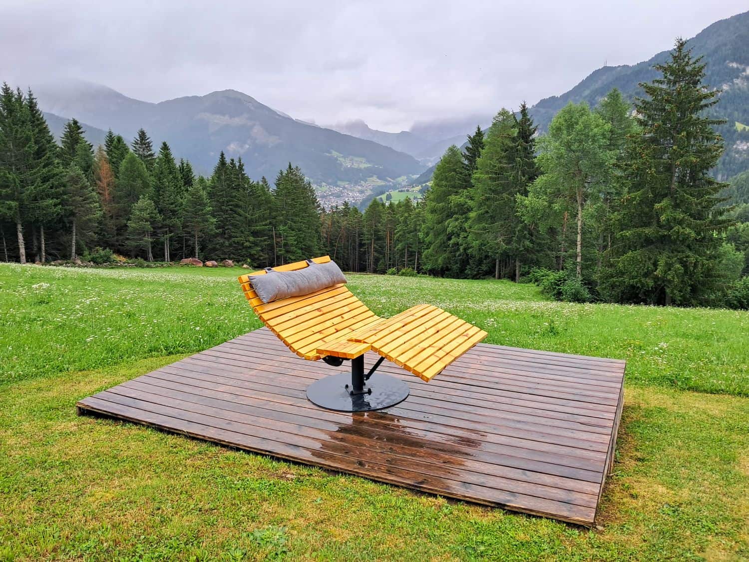 A modern wooden lounge chair with a cushion sits on a wet wooden platform in a grassy field, surrounded by pine trees and mountains under a cloudy sky.